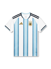 アルゼンチン代表2026ホームジャージ正面、ホワイトとライトブルーの縦ストライプ、ゴールド調の胸ロゴ、三つ星付き代表エンブレム、チャンピオンバッジなし、ネイビーの肩3ストライプ、半袖デザイン