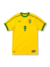 イエローベースにグリーンの襟と肩口ラインを配し、胸にナイキロゴ、星4つ付きCBFブラジルサッカー連盟エンブレムとBRASIL表記、ブルーの9番をあしらったブラジル代表1998ワールドカップホームジャージ正面