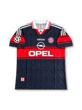 バイエルン・ミュンヘン 1997〜1999年 アウェイユニフォーム ローター・マテウス10番。adidas製のダークネイビーのボディにレッドの太い胸ラインと袖の切り替え、ホワイトの折り襟と肩のスリーストライプを組み合わせたレトロサッカージャージ。胸元にはadidasロゴ、バイエルンのクラブエンブレム、ホワイトのOPELスポンサーロゴを配置。UEFAチャンピオンズリーグの袖章を備え、背面にはMATTHÄUSのネームとホワイトの10番を配したデザイン。