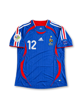 フランス代表 2006年ドイツワールドカップ ホームユニフォーム ティエリ・アンリ12番。adidas製ロイヤルブルーのボディに細かな縦ラインの織り柄、レッドの襟元、ホワイトの肩のスリーストライプとレッドの曲線状サイドパネルを組み合わせたレトロサッカージャージ。胸元にはホワイトのadidasロゴ、ゴールドスター付きフランス代表エンブレム、ホワイトの12番を配置。袖には2006年ワールドカップの大会デザインパッチを備え、背面にはHENRYのネームとホワイトの12番を配したデザイン。