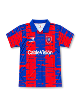 サン・ロレンソ 1990年代 グラフィックユニフォーム。Penalty製のレッドとネイビーブルーを組み合わせたボディ全面に、大胆な幾何学模様と大きな文字を重ねた1995/96シーズンを思わせるレトロサッカージャージ。ネイビーの折り返し襟を採用し、胸元にはホワイトのPenaltyロゴ、サン・ロレンソのクラブエンブレム、CableVisiónスポンサーロゴを配置。伝統的な縦ストライプとは異なる、1990年代のアルゼンチンサッカーらしい個性的な総柄デザイン。
