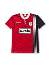 Oasis × adidas Originals Live '25 ツアー マンチェスタージャージ レッド 深みのあるカーディナルレッドとブラックを左右非対称に切り替えたデザイン、ホワイトのヘンリーネックカラー、adidas Originalsのスリーストライプス、ホワイトのadidasロゴ、中央のOasisボックスロゴ、マンチェスターのパブBoltmakers Armsをモチーフにしたシールドエンブレムを配したツアー記念フットボールジャージ
