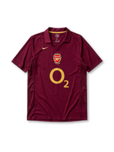 レッドカラント地にゴールドパイピングのポロ襟、左胸のゴールドNikeスウッシュ、中央のHIGHBURY 1913-2006記念エンブレム、大きなO2スポンサーロゴ、両脇のメッシュ通気パネルを備えたアーセナル2005-06ホームジャージ正面