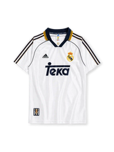 ホワイトベースにネイビー・ゴールドのスリーストライプ、REAL MADRIDロゴ入りのポロ襟、左胸のadidasロゴ、右胸のレアル・マドリードクラブエンブレム、中央のネイビーのTekaスポンサーロゴを備えたレアル・マドリード1998/00ホームジャージ正面